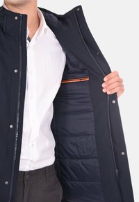 Giacca imbottita blu navy con chiusura a zip, bottoni a scatto e tasca interna con accenti arancioni; superficie liscia con foderatura trapuntata.