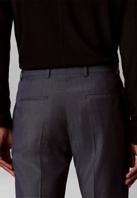 Vue arrière d'une personne portant un pantalon habillé gris foncé avec passants de ceinture et poches arrière passepoilées, associé à un haut noir à manches longues.