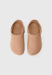Ett par beige slip-on Crocs med texturerade ovandelar och justerbara hälremmar, visade ovanifrån på en vit bakgrund.
