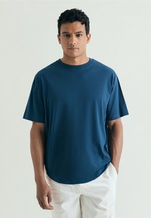 Man standing against plain light background, wearing a blue short-sleeve t-shirt and white pants, hands in pockets, facing forward.
