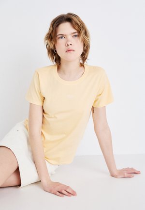 T-shirt en coton jaune pâle avec un col rond, des manches courtes et un logo discret sur la poitrine, associé à une jupe blanche.