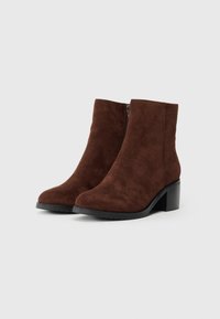 Braune Wildleder-Stiefeletten mit runder Zehenform, seitlichem Reißverschluss und einem gestapelten schwarzen Absatz. Glatte Textur ohne sichtbare Muster oder Akzente.