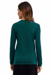 Donna con lunghi capelli castani che indossa una maglietta fitted a maniche lunghe verde scuro, di spalle contro uno sfondo bianco.