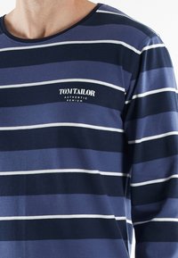 Chemise à manches longues bleu marine avec des rayures blanches horizontales et un col rond. Présente le logo "TOM TAILOR" en blanc sur la poitrine. Texture de tissu douce.