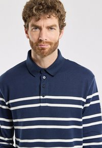 Polo bleu marine avec des rayures horizontales blanches. En coton, il présente un col et une patte à trois boutons. Tissu en maille texturée.