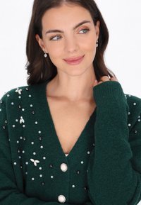 Zielony kardigan z dekoltem w serek, ozdobiony różnymi koralikami i kwiatowymi akcentami. Posiada duże dekoracyjne guziki i ściągacze przy mankietach.
