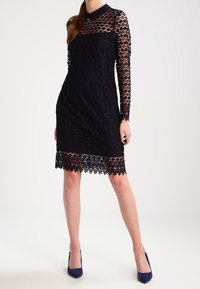 Schwarzes Spitzenkleid mit langen Ärmeln und einem Kragen, ausgestattet mit einem Blumendesign und einem gewellten Saum. Getragen mit marineblauen Absatzschuhen.