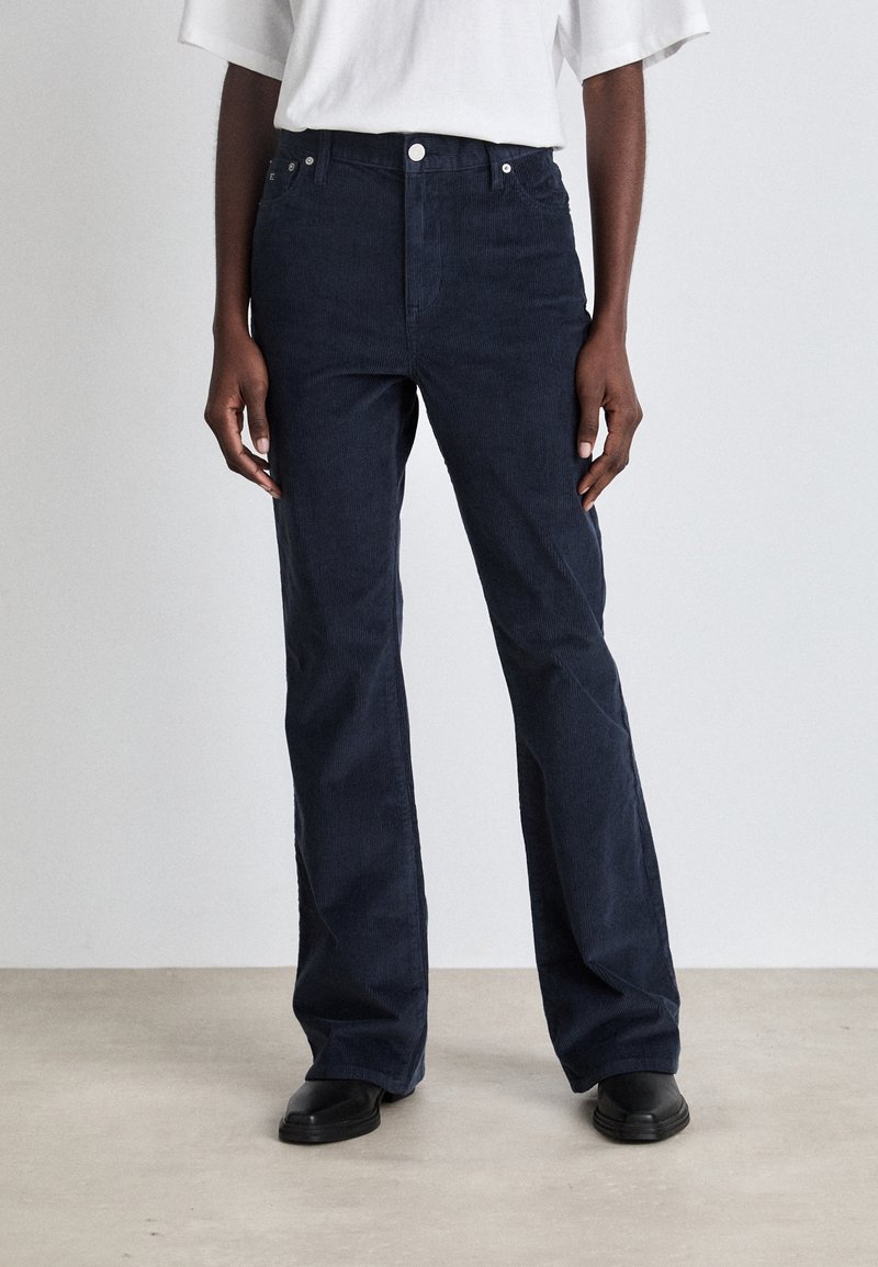 Pantalon en velours côtelé bleu marine avec taille haute, jambes droites, poches avant, fermeture à bouton en métal et surface texturée. Associé à des chaussures noires.