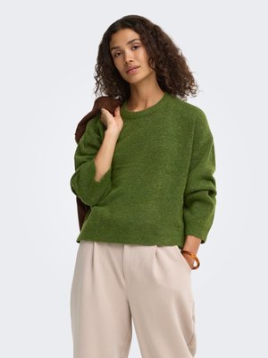 Femme aux cheveux bouclés foncés portant un pull vert et un pantalon beige, tenant une veste marron sur son épaule devant un fond uni.