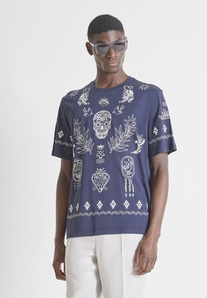 Hombre con camiseta azul marino con calavera blanca del Día de los Muertos y diseños botánicos, pantalones blancos y grandes gafas de sol grises.