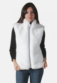 Gilet en fausse fourrure blanche avec un col haut et une fermeture éclair sur toute la longueur. Il présente une texture douce et un design sans manches, porté par-dessus un haut noir à manches longues.