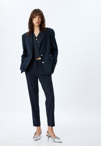 Navy maßgeschneiderter Blazer mit silbernen Knöpfen und passenden schmal geschnittenen Hosen, kombiniert mit metallisch silbernen Absatzschuhen. Strukturiertes Gewebe.
