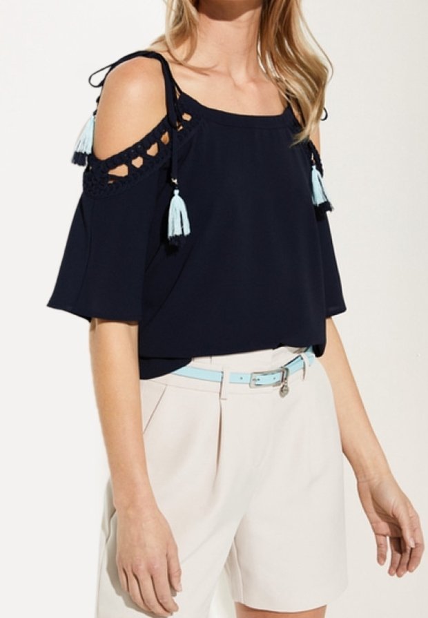 Marineblaue Cold-Shoulder-Bluse mit gehäkelten Akzenten und hellblauen Quasten; getragen mit cremefarbenen Shorts und einem passenden hellblauen Gürtel.