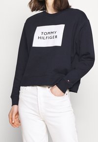 Donna che indossa un felpa nera corta con una patch bianca del logo "Tommy Hilfiger" e jeans bianchi a vita alta, con una mano in tasca.