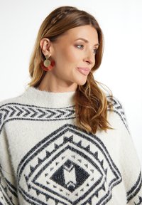 Orecchini circolari in oro con frange e pon pon rossi, design testurizzato. Il maglione presenta una base crema con pattern geometrici neri.