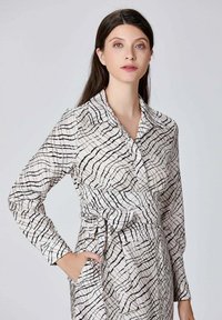Robe à motif noir et blanc avec un col, des manches longues, une ceinture à nouer à la taille et deux poches latérales. Tissu à texture lisse.