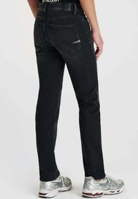 Jean en denim noir avec une coupe ajustée, comportant deux poches arrière et une ceinture texturée. Associé à des chaussures de sport argentées.
