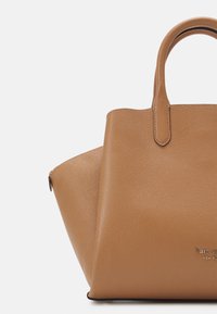 Brązowa skórzana torba tote z podwójnymi uchwytami, trójkątnymi bocznymi panelami i logo "kate spade new york" w złotym kolorze.