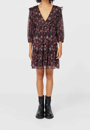 Vrouw draagt een zwarte, bloemenprint knielange jurk met driekwartmouwen en een diepe V-hals, gecombineerd met zwarte enkellaarsjes.