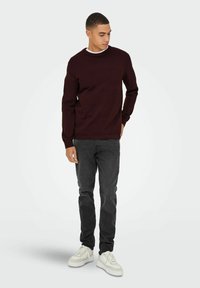 Bordeaux gekleurde textuurtrui met ronde hals, gecombineerd met zwarte jeans en witte sneakers. Model staat casual met één hand in de zak.