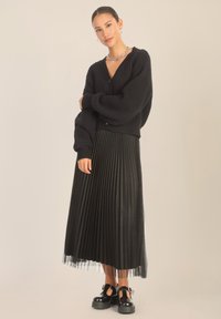 Czarny kardigan z ribbed w serek i zapięciem na guziki, w połączeniu z czarną spódnicą plisowaną z przezroczystym akcentem przy dolnej krawędzi. Masywne czarne buty.