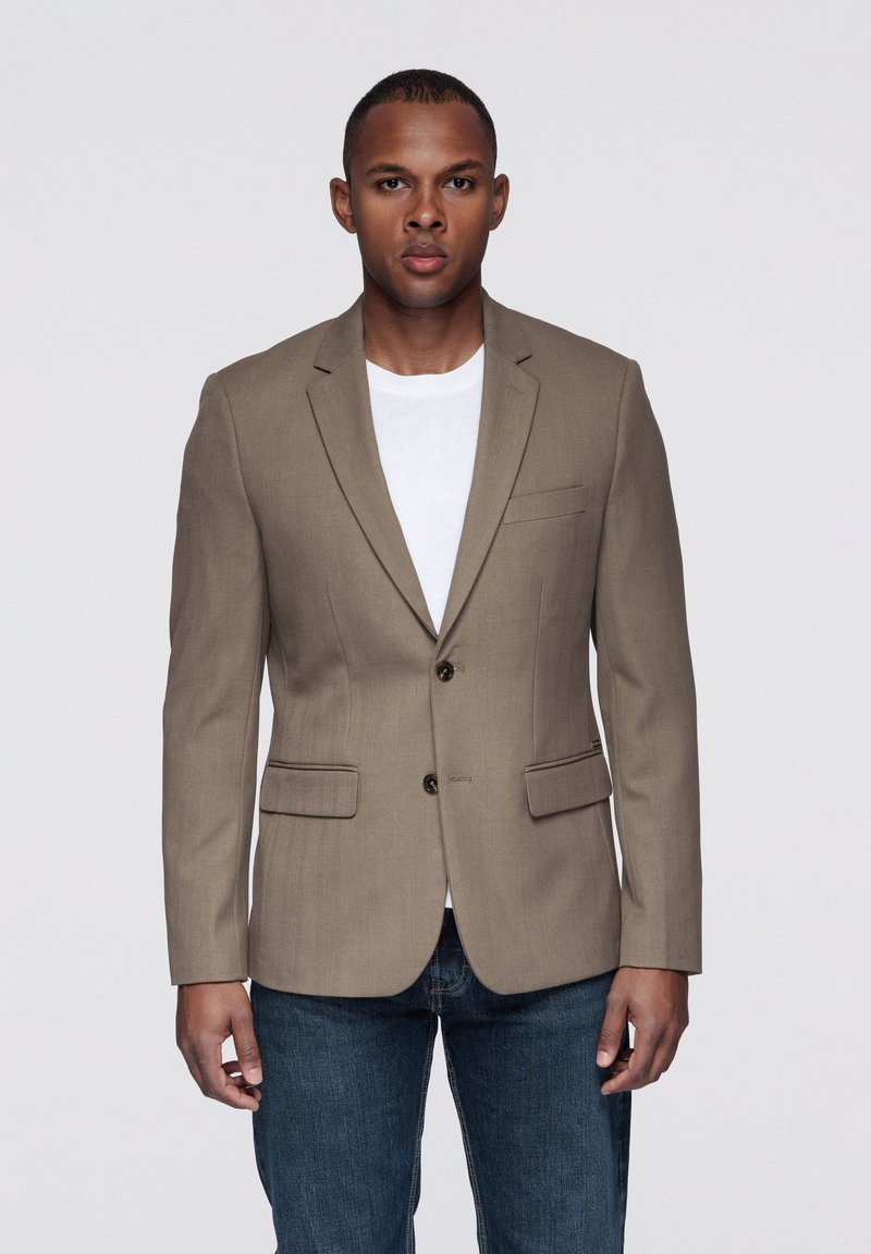 Beżowa marynarka o dopasowanym kroju, z dwoma przednimi kieszeniami i trzema guzikami. Noszona na białej koszuli i zestawiona z ciemnoniebieskimi dżinsami.