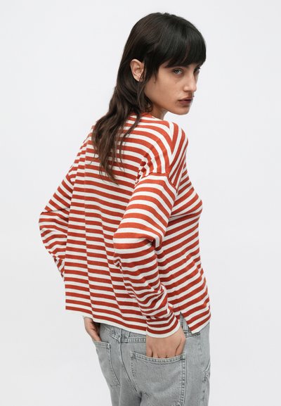 Πουλόβερ με κόκκινες και λευκές ριγέ, μακριά μανίκια, άνετη εφαρμογή και πλευρικές ανοίγματα, φορεμένο πάνω από ανοιχτόχρωμα γκρι τζιν. Ορατό υφή πλεκτού ύφασματος.