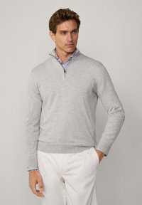 Grauer Pullover aus weichem Material, mit einem Reißverschluss-Kragen und gerippten Bündchen. Getragen über einem blau-weiß gestreiften Hemd, kombiniert mit weißen Hosen.