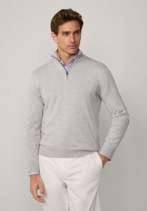 Grauer Pullover aus weichem Material, mit einem Reißverschluss-Kragen und gerippten Bündchen. Getragen über einem blau-weiß gestreiften Hemd, kombiniert mit weißen Hosen.