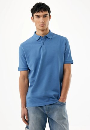 Junger Mann trägt ein blaues kurzärmeliges Poloshirt mit Kragen und hellblaue Jeans, steht mit einer Hand in der Tasche.