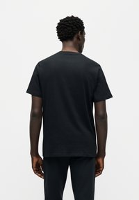 Μαύρο βαμβακερό μπλουζάκι με κλασικό στρογγυλό λαιμόκοψη και κοντά μανίκια, εμφανιζόμενο από την πίσω πλευρά. Ομαλή υφή και άνετη εφαρμογή.