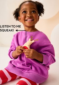 Paarse sweatshirt met geribbelde manchetten, rood-roze gestreepte legging, houdend een speelgoed rendier met een pieper. Achtergrond met zachte, neutrale tinten.