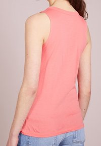 Top in corallo senza maniche realizzato in tessuto morbido, con una silhouette aderente, scollatura rotonda e una consistenza liscia, abbinato a jeans blu.