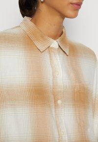 Camisa de botones con un patrón a cuadros en tonos beige claro y crema, con una textura de tela suave y un cuello clásico. Botones sutiles visibles.