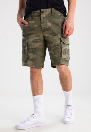 Homme portant un short cargo camouflage vert, des chaussettes blanches mi-mollet avec des logos, et des baskets blanches Adidas, debout les mains près des poches.