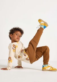 Langärmliges cremefarbenes Hemd mit Löwenmotiven, kombiniert mit braunen Sweatpants mit strukturierten Akzenten. Trägt gelbe Sneakers mit blauen Details.
