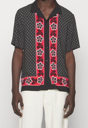 Schwarzes Kurzarmhemd mit kleinen weißen Mustern und vertikalen roten Einsätzen mit großen weißen und schwarzen Blumen und Blättern, getragen zu weißen Hosen.