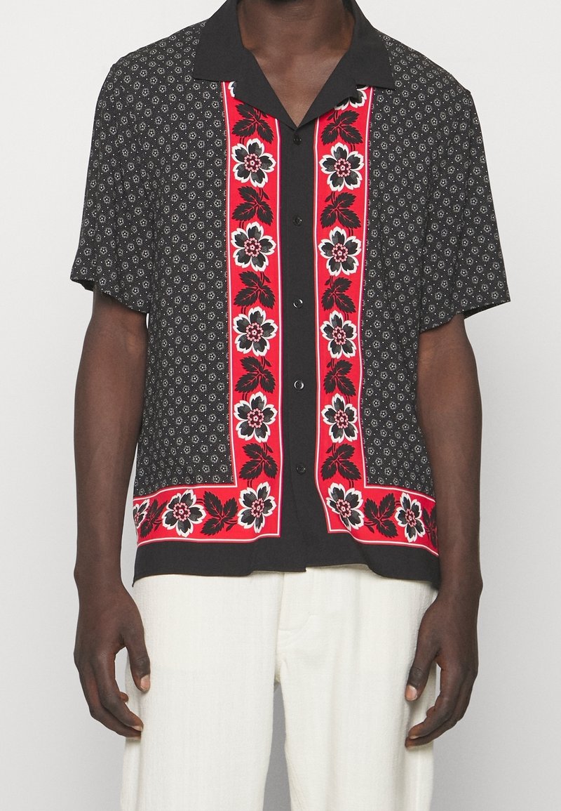 Chemise noire à manches courtes avec petits motifs blancs et panneaux verticaux rouges présentant de grandes fleurs blanches et noires ainsi que des feuilles, portée avec un pantalon blanc.