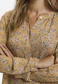 Gelbe Bluse mit floralen Mustern, die kleine rosa und schwarze Designs zeigt, mit einem Knopfverschluß am Hals und langen Ärmeln mit gerafften Bündchen. Strukturierter Stoff.