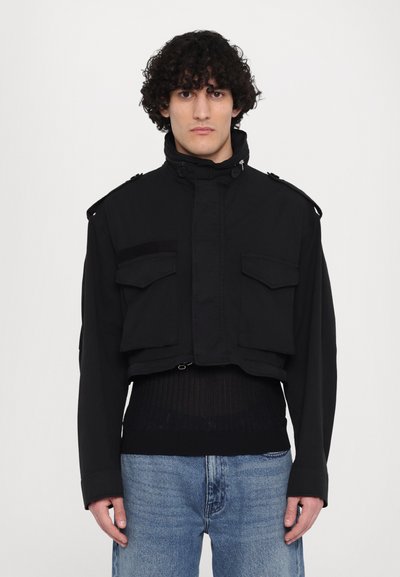 Zwarte cropped jas met schouderepauletten, twee voorzakken en een hoge kraag. Gemaakt van een gladde stof met een matte afwerking.