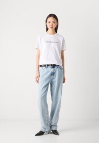 Balta medvilninė marškinė su "Calvin Klein Jeans" logotipu, derinama su šviesiai mėlynomis džinsinėmis kelnėmis ir juodais smailaus priekio aukštakulniais bateliais.