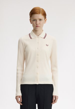 Ung kvinde med kort rødt hår iført en cremefarvet ribbet langærmet skjorte med knapper, med en krave med mørkerøde striber, og sorte bukser.