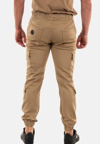 Beige cargobroek met elastische tailleband, meerdere zijzakken en opgerolde enkels. Gladde stoftextuur, voorzien van een patch op de achterkant.
