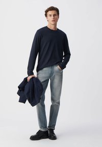 Navy-langärmliges Shirt, helle Denim-Jeans und schwarze Schuhe. Das Modell hält eine marinefarbene Jacke. Glattes Material mit entspannter Passform und geradem Bein.