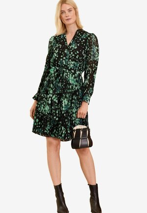 Schwarzes Kleid mit einem grünen abstrakten Muster, langen Ärmeln, einer knöpfbaren Front und einem taillierten Bund. Kombiniert mit einer kleinen schwarzen Handtasche und Stiefeln bis zum Knöchel.