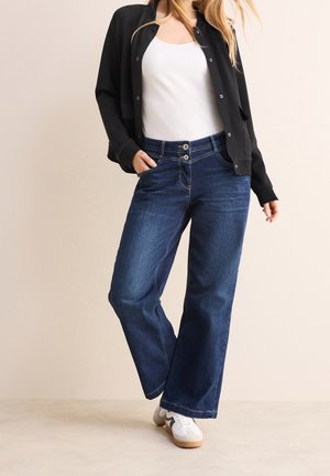 Kobieta ubrana w czarną kurtkę na guziki, biały tank top, szerokie niebieskie spodnie dżinsowe i białe trampki, pozująca z jedną ręką w kieszeni.