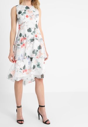 Robe blanche fleurie sans manches avec un ourlet asymétrique en couches, portée avec des sandales noires à talons hauts et bride à la cheville.