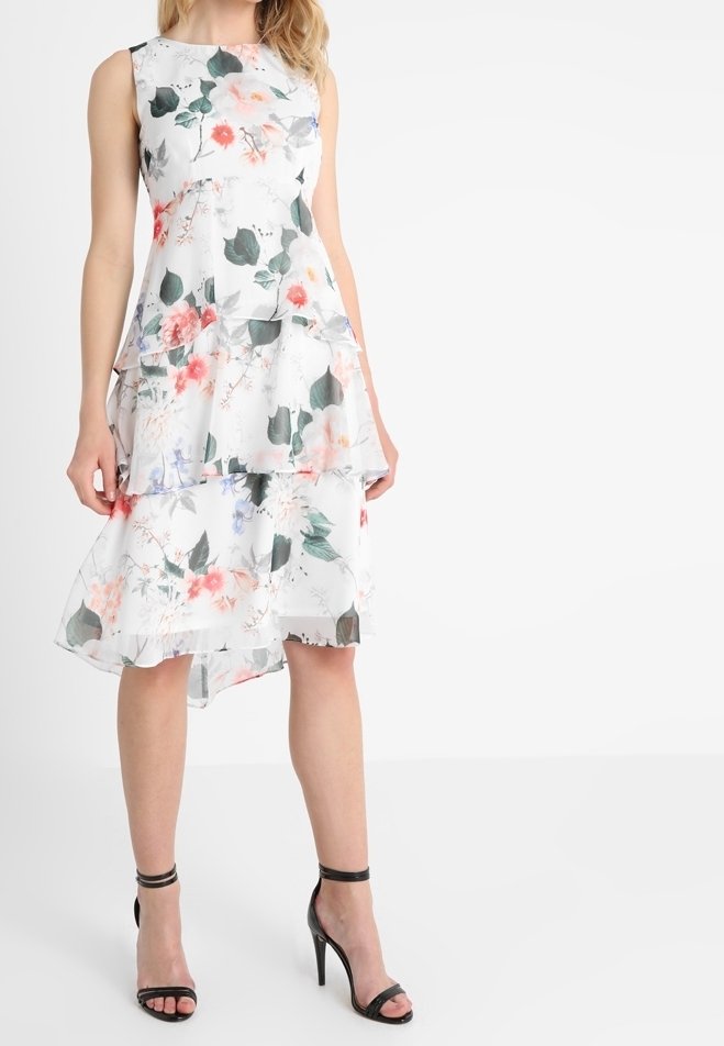 Robe blanche fleurie sans manches avec un ourlet asymétrique en couches, portée avec des sandales noires à talons hauts et bride à la cheville.