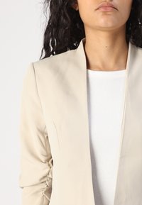 Jasno beżowa marynarka o dopasowanym kroju, z kołnierzem szalowym i marszczonymi rękawami, nałożona na białą koszulkę z okrągłym dekoltem.