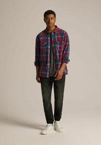 Czerwona i niebieska koszula flanelowa w kratę, szary t-shirt pod spodem, czarne dżinsy i białe sneakersy, stojący na neutralnym tle.